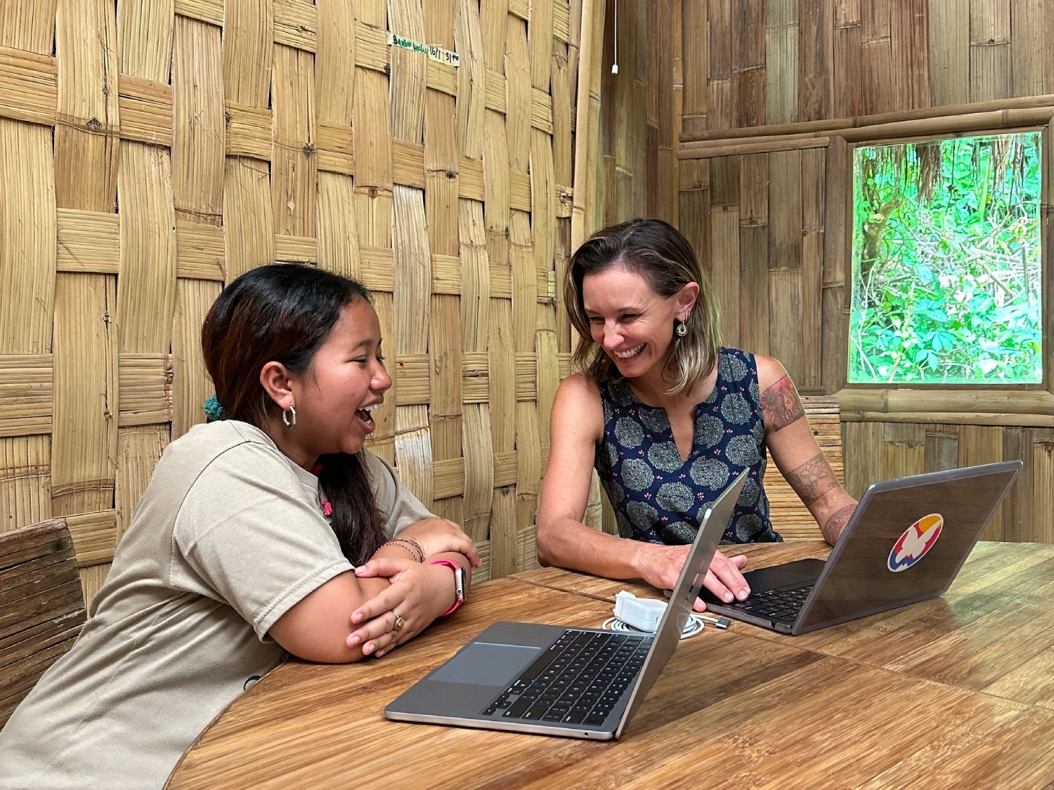 Two women smiling with laptops in bamboo hut